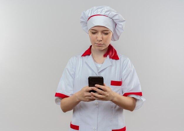 Il giovane chef femminile biondo fiducioso in uniforme del cuoco unico tiene ed esamina il telefono isolato sulla parete bianca