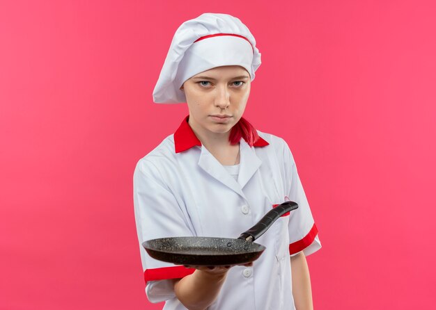 Il giovane chef femminile biondo fiducioso in uniforme del cuoco unico tiene la padella e sembra isolato sulla parete rosa
