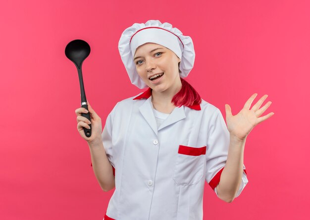 Il giovane chef femminile biondo gioioso in uniforme del cuoco unico tiene il mestolo e tiene la mano aperta isolata sulla parete rosa