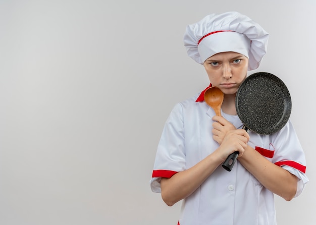 Il giovane chef femminile biondo infastidito in uniforme del cuoco unico attraversa le braccia e tiene la padella e il cucchiaio isolati sulla parete bianca