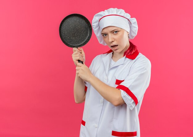 Il giovane chef femminile biondo infastidito in uniforme del cuoco unico tiene la padella con entrambe le mani isolate sulla parete rosa