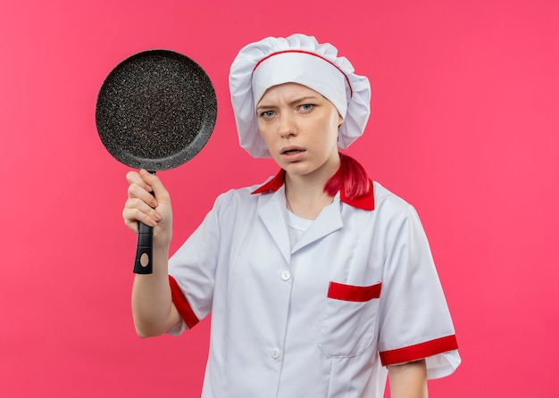 Il giovane chef femminile biondo infastidito in uniforme del cuoco unico tiene la padella e sembra isolato sulla parete rosa