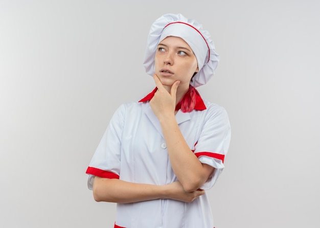 Il giovane chef femminile biondo premuroso in uniforme del cuoco unico mette la mano sul mento isolato sulla parete bianca