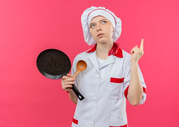Il giovane chef femminile biondo premuroso in uniforme dello chef tiene la padella e il cucchiaio di legno rivolto verso l'alto isolato sulla parete rosa