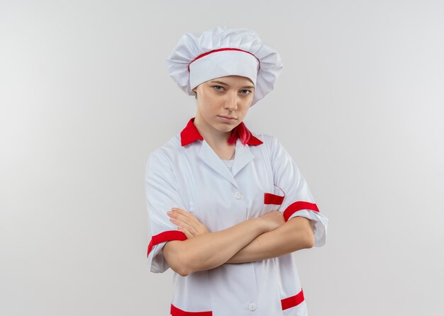 Il giovane chef femminile biondo sicuro in uniforme del cuoco unico attraversa le braccia e sembra isolato sulla parete bianca