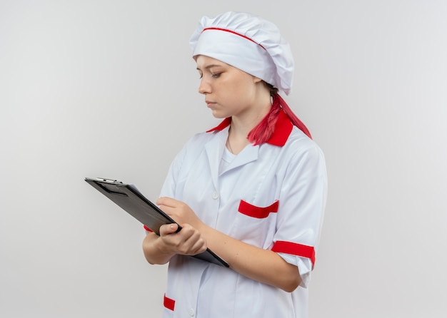 Il giovane chef femminile biondo sicuro in uniforme del cuoco unico tiene ed esamina la lavagna per appunti isolata sulla parete bianca