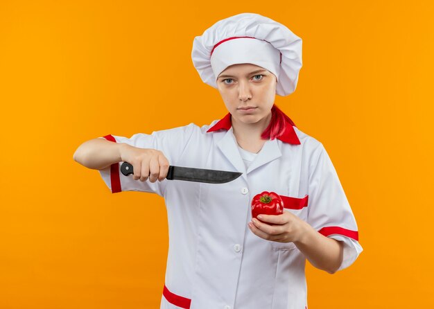 Il giovane chef femminile biondo sicuro in uniforme del cuoco unico tiene il coltello e il peperone rosso isolato sulla parete arancione