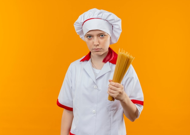 Il giovane chef femminile biondo sicuro in uniforme del cuoco unico tiene il mazzo di spaghetti isolato sulla parete arancione