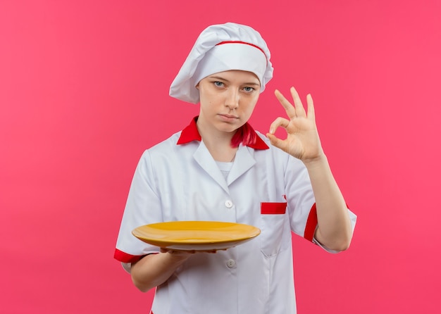 Il giovane chef femminile biondo sicuro in uniforme del cuoco unico tiene il segno giusto della mano di gesti e del piatto isolato sulla parete rosa