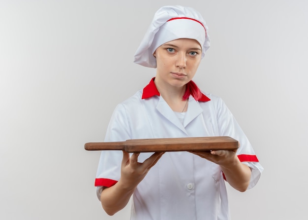 Il giovane chef femminile biondo sicuro in uniforme del cuoco unico tiene il tagliere isolato sulla parete bianca