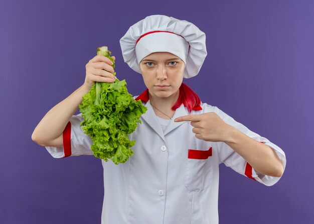Il giovane chef femminile biondo sicuro in uniforme del cuoco unico tiene l'insalata capovolta e punti con il dito isolato sulla parete viola