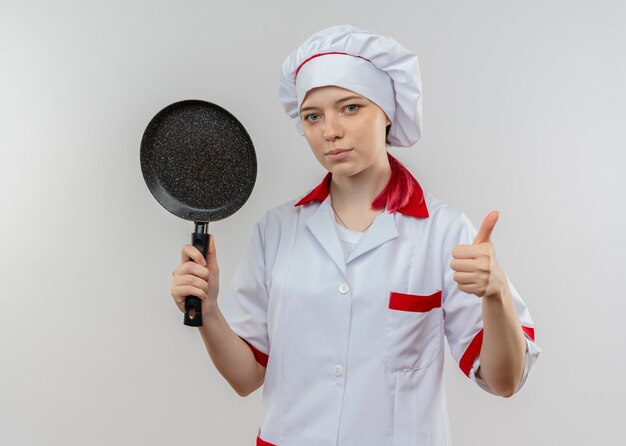 Il giovane chef femminile biondo sicuro in uniforme del cuoco unico tiene la padella ed i pollici in su isolati sulla parete bianca