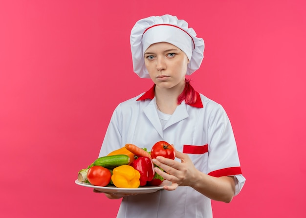 Il giovane chef femminile biondo sicuro in uniforme del cuoco unico tiene le verdure sulla zolla e sembra isolato sulla parete rosa