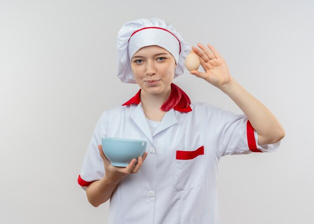 Il giovane chef femminile biondo soddisfatto in uniforme del cuoco unico tiene la ciotola e l'uovo isolati sulla parete bianca