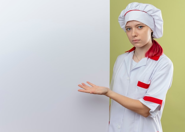 Il giovane chef femminile biondo sorpreso in uniforme da chef sta dietro e indica il muro bianco che sembra isolato sulla parete verde