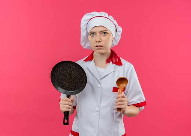 Il giovane chef femminile biondo sorpreso in uniforme da chef tiene la padella e il cucchiaio di legno isolato sulla parete rosa