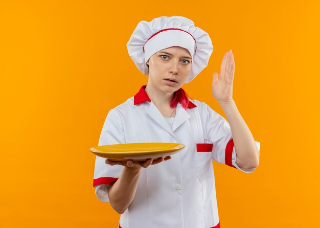 Il giovane chef femminile biondo sorpreso in uniforme del cuoco unico tiene il piatto e solleva la mano isolata sulla parete arancione