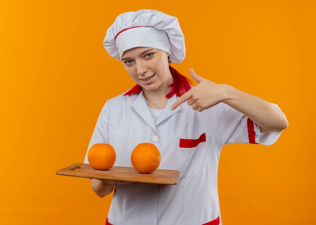 Il giovane chef femminile biondo sorridente in uniforme del cuoco unico tiene le arance sul tagliere e punti con il dito isolato sulla parete arancio