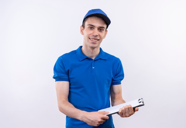Il giovane corriere che indossa l'uniforme blu e il berretto blu sorride e scrive su una lavagna per appunti