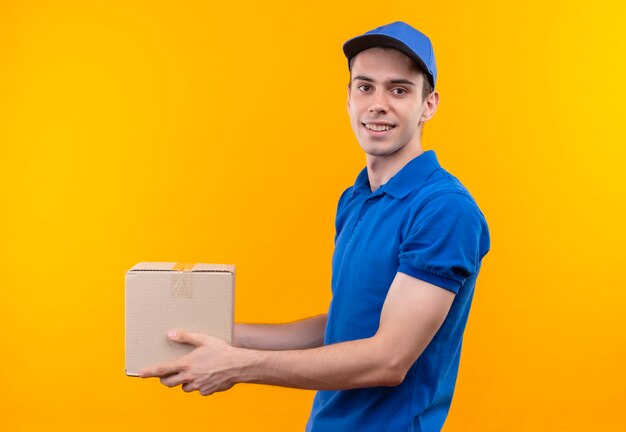 Il giovane corriere che indossa l'uniforme blu e il berretto blu sorride e tiene la scatola