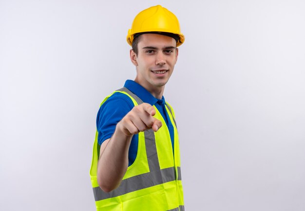 Il giovane costruttore che indossa l'uniforme da costruzione e il casco di sicurezza sorride e ti indica con l'indice