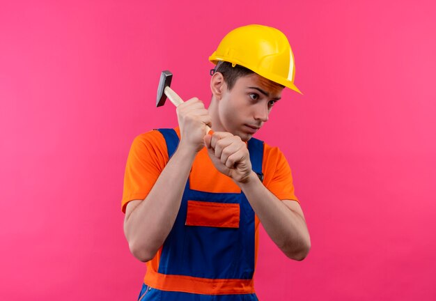 Il giovane costruttore che indossa l'uniforme da costruzione e il casco di sicurezza tiene un martello