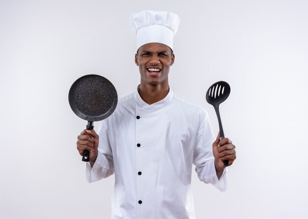 Il giovane cuoco afro-americano infastidito in uniforme dello chef tiene la padella e la spatola su fondo bianco isolato con lo spazio della copia