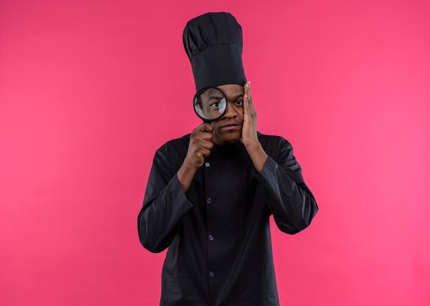 Il giovane cuoco afroamericano ansioso in uniforme del cuoco unico osserva tramite la lente d'ingrandimento o la lente di ingrandimento isolata su fondo rosa con lo spazio della copia