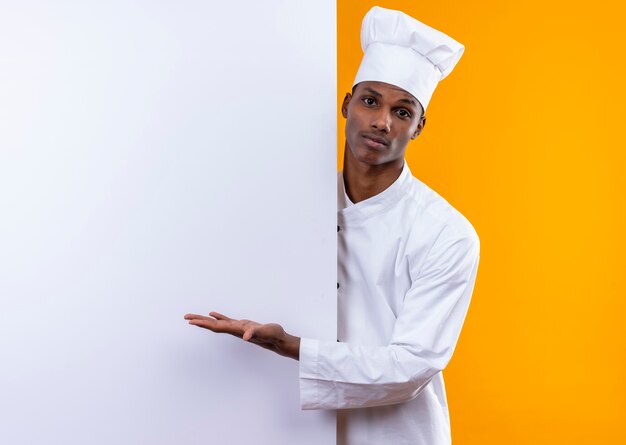 Il giovane cuoco afroamericano confuso in uniforme del cuoco unico sta dietro la parete bianca e indica alla parete con la mano isolata sulla parete arancione