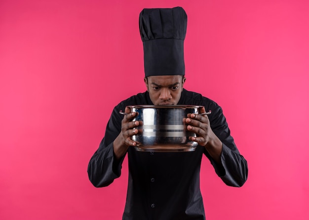 Il giovane cuoco afroamericano confuso in uniforme del cuoco unico tiene ed esamina la casseruola isolata sulla parete rosa