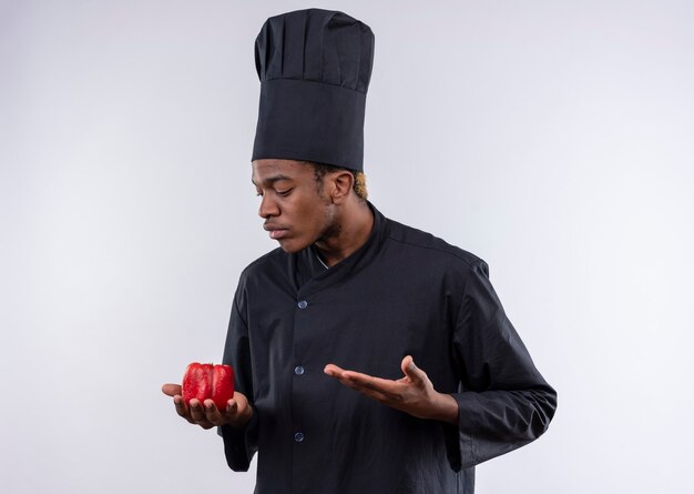 Il giovane cuoco afroamericano confuso in uniforme del cuoco unico tiene il peperone rosso e punti con la mano isolata sulla parete bianca