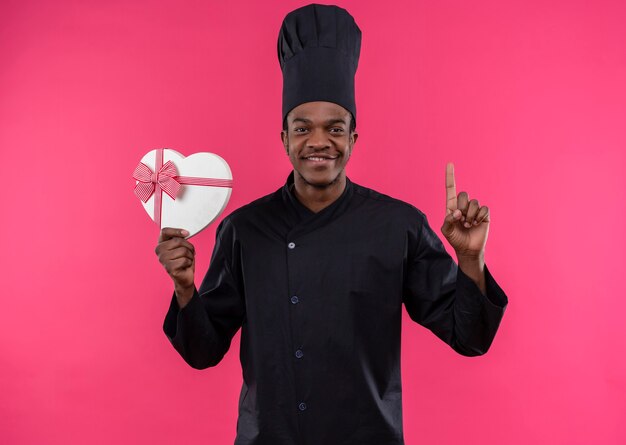 Il giovane cuoco afroamericano contento in uniforme dello chef tiene la scatola a forma di cuore e il pollice in alto isolato sulla parete rosa