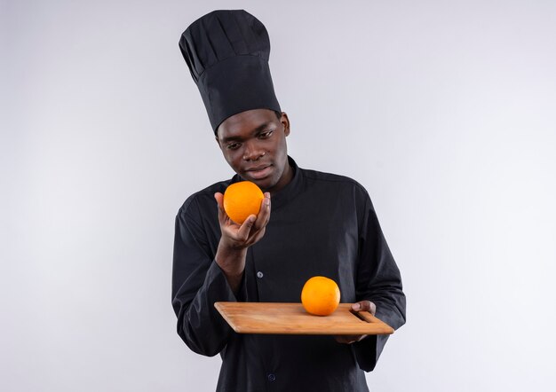 Il giovane cuoco afroamericano felice in uniforme del cuoco unico tiene l'arancia sul tagliere e sulla mano su bianco con lo spazio della copia