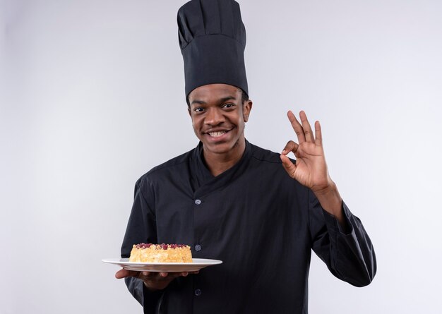 Il giovane cuoco afroamericano felice in uniforme dello chef tiene la torta sul piatto e gesti il segno giusto della mano isolato sulla parete bianca