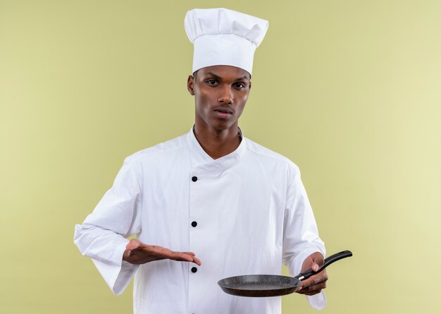 Il giovane cuoco afroamericano fiducioso in uniforme da chef tiene la padella e punti con la mano isolata su sfondo verde con spazio di copia