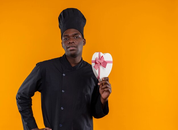 Il giovane cuoco afroamericano fiducioso in uniforme da chef tiene la scatola a forma di cuore e guarda la telecamera sull'arancio con lo spazio della copia