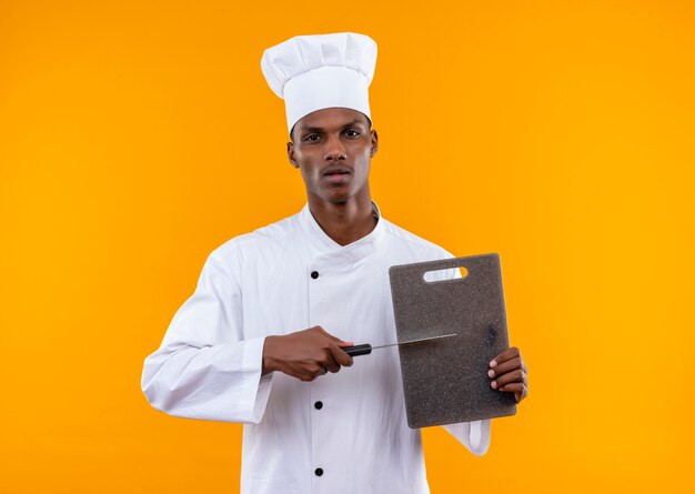 Il giovane cuoco afroamericano fiducioso in uniforme da chef tiene lo scrittorio della cucina e il coltello isolato su priorità bassa arancione con lo spazio della copia