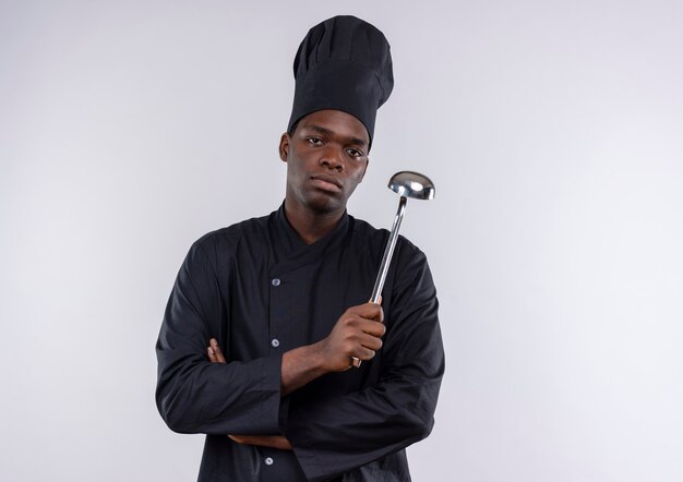 Il giovane cuoco afroamericano fiducioso in uniforme del cuoco unico attraversa le braccia tiene il mestolo su bianco con lo spazio della copia