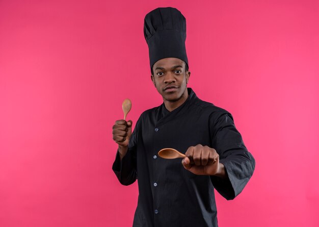 Il giovane cuoco afroamericano fiducioso in uniforme dello chef tiene i cucchiai di legno isolati su fondo rosa con lo spazio della copia