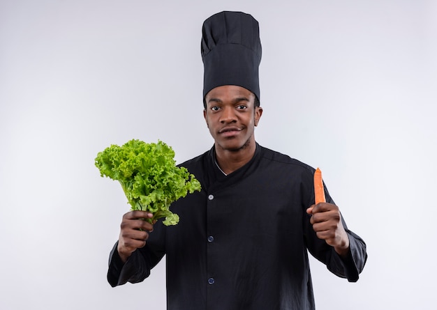Il giovane cuoco afroamericano fiducioso in uniforme dello chef tiene insalata verde e carota isolato su priorità bassa bianca con lo spazio della copia