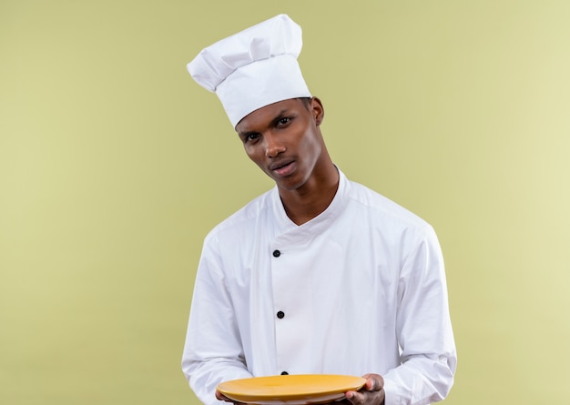 Il giovane cuoco afroamericano fiducioso in uniforme dello chef tiene la piastra con entrambe le mani isolato su sfondo verde con spazio di copia