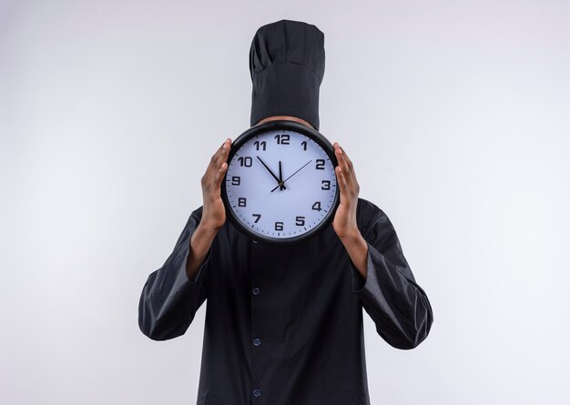 Il giovane cuoco afroamericano in uniforme del cuoco unico chiude il fronte con l'orologio isolato su fondo bianco con lo spazio della copia