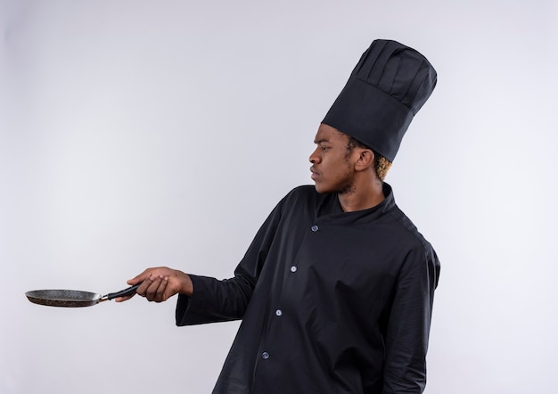 Il giovane cuoco afroamericano infastidito in uniforme dello chef sta lateralmente e tiene la padella isolata su fondo bianco con lo spazio della copia