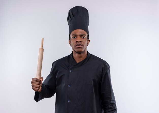 Il giovane cuoco afroamericano infastidito in uniforme dello chef tiene il mattarello isolato su priorità bassa bianca con lo spazio della copia