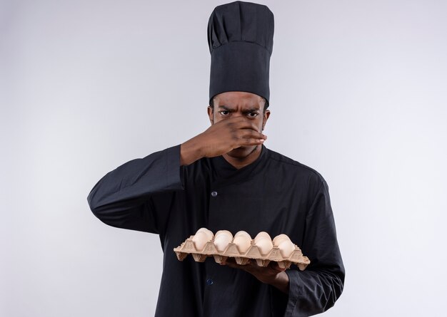 Il giovane cuoco afroamericano infastidito in uniforme dello chef tiene un lotto di uova e chiude il naso con la mano isolata su fondo bianco con lo spazio della copia