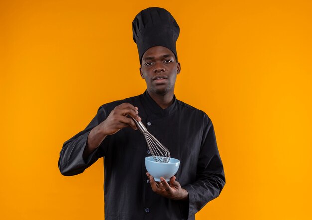 Il giovane cuoco afroamericano sicuro in uniforme del cuoco unico tiene la frusta e la ciotola sull'arancia con lo spazio della copia