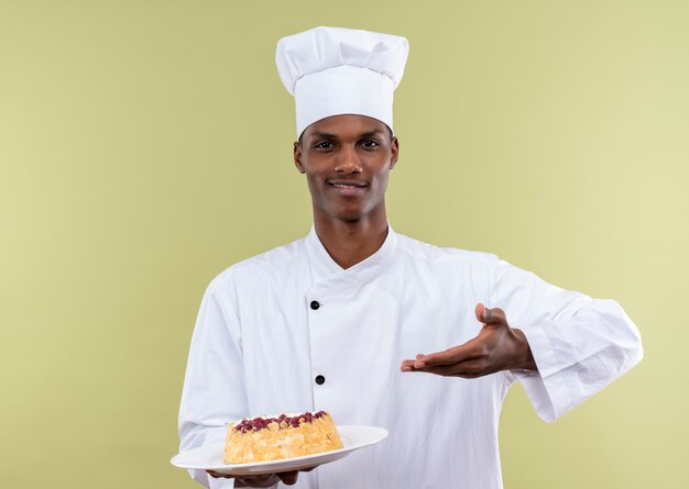 Il giovane cuoco afroamericano soddisfatto in uniforme del cuoco unico indica alla torta sulla zolla bianca e sui punti con la mano isolata sulla parete verde