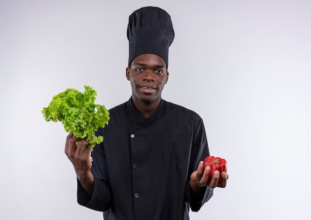 Il giovane cuoco afroamericano soddisfatto in uniforme del cuoco unico tiene insalata e peperone rosso su bianco con lo spazio della copia