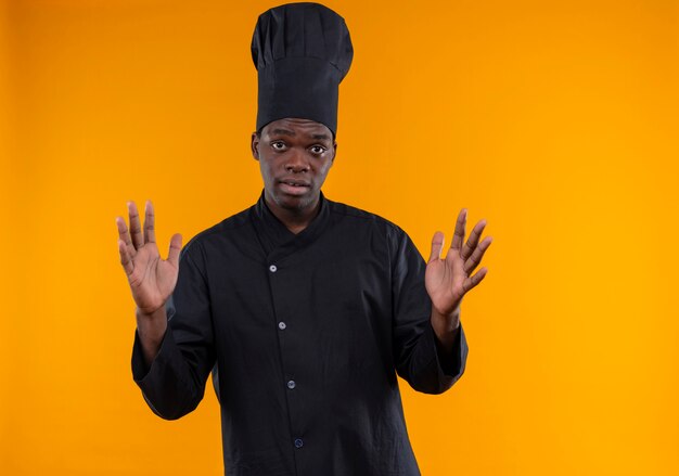 Il giovane cuoco afroamericano sorpreso in uniforme dello chef tiene le mani in alto guardando la telecamera sull'arancio con lo spazio della copia