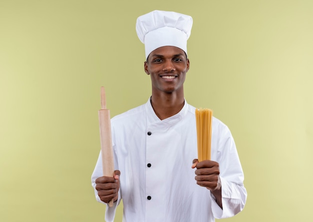 Il giovane cuoco afroamericano sorridente in uniforme del cuoco unico tiene il mattarello e il mazzo di spaghetti nelle mani isolate sulla parete verde